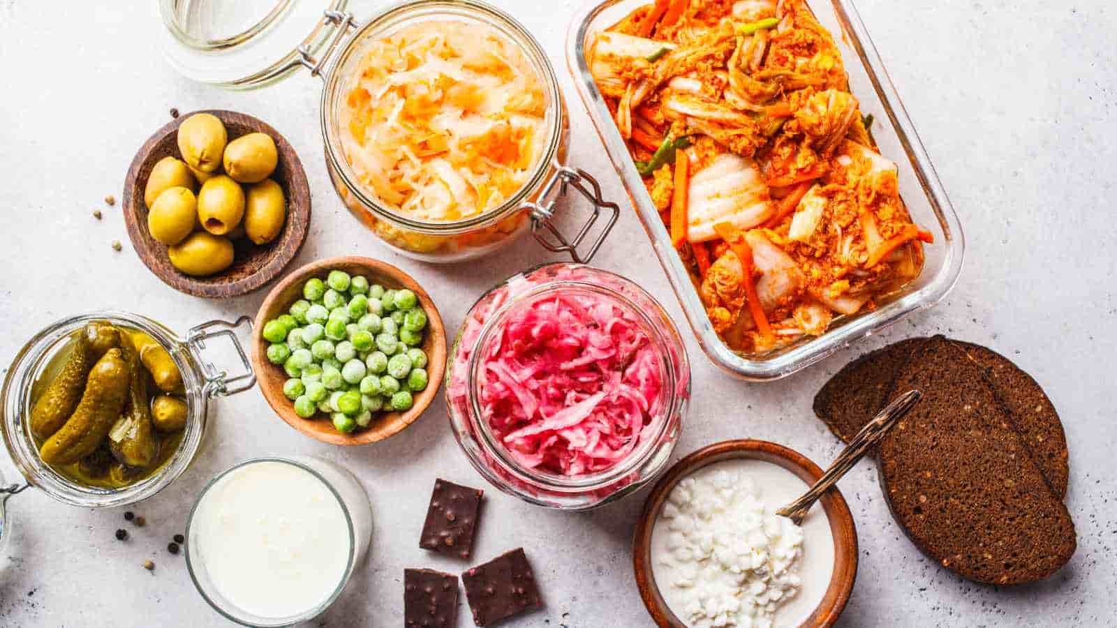Assorted fermented foods including kimchi, sauerkraut, pickles, olives, and cottage cheese arranged on a light surface with bread, crackers, and a small bowl of frozen peas.