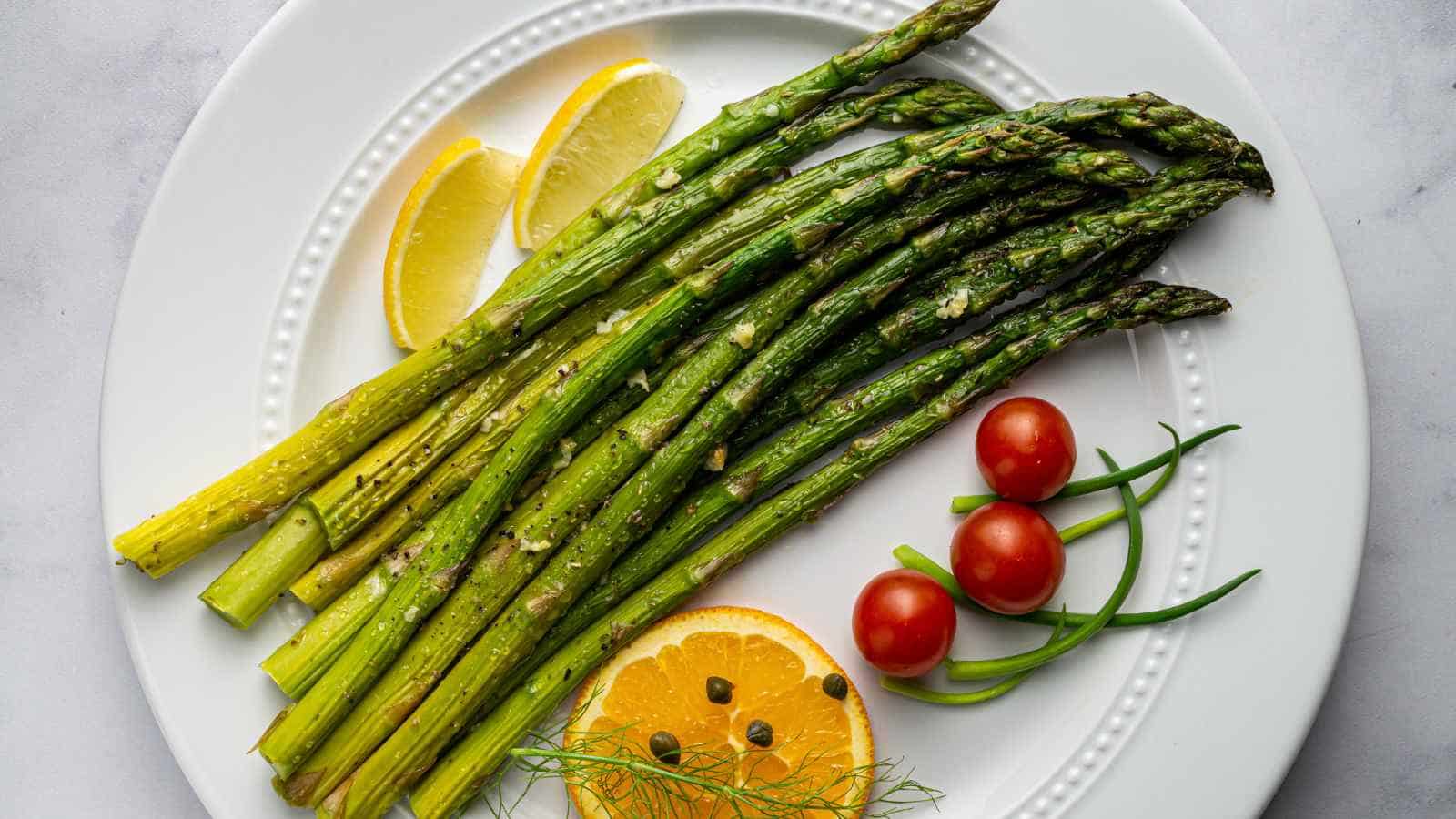Grilled asparagus on a white plate with lemon slices, cherry tomatoes, orange slices, capers, and green garnish.
