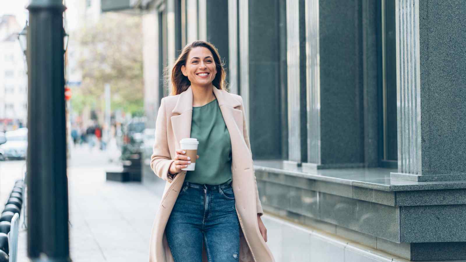 A woman in a beige coat and green top walks down a city street holding a coffee cup, smiling, with buildings and pedestrians in the background.