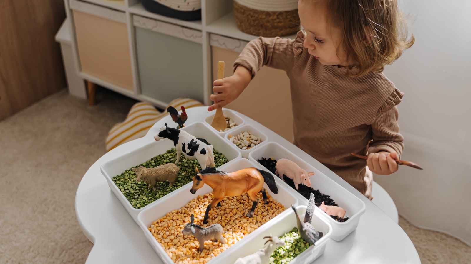 Child playing with farm animal toys in a sensory bin filled with dried beans and peas on a table.