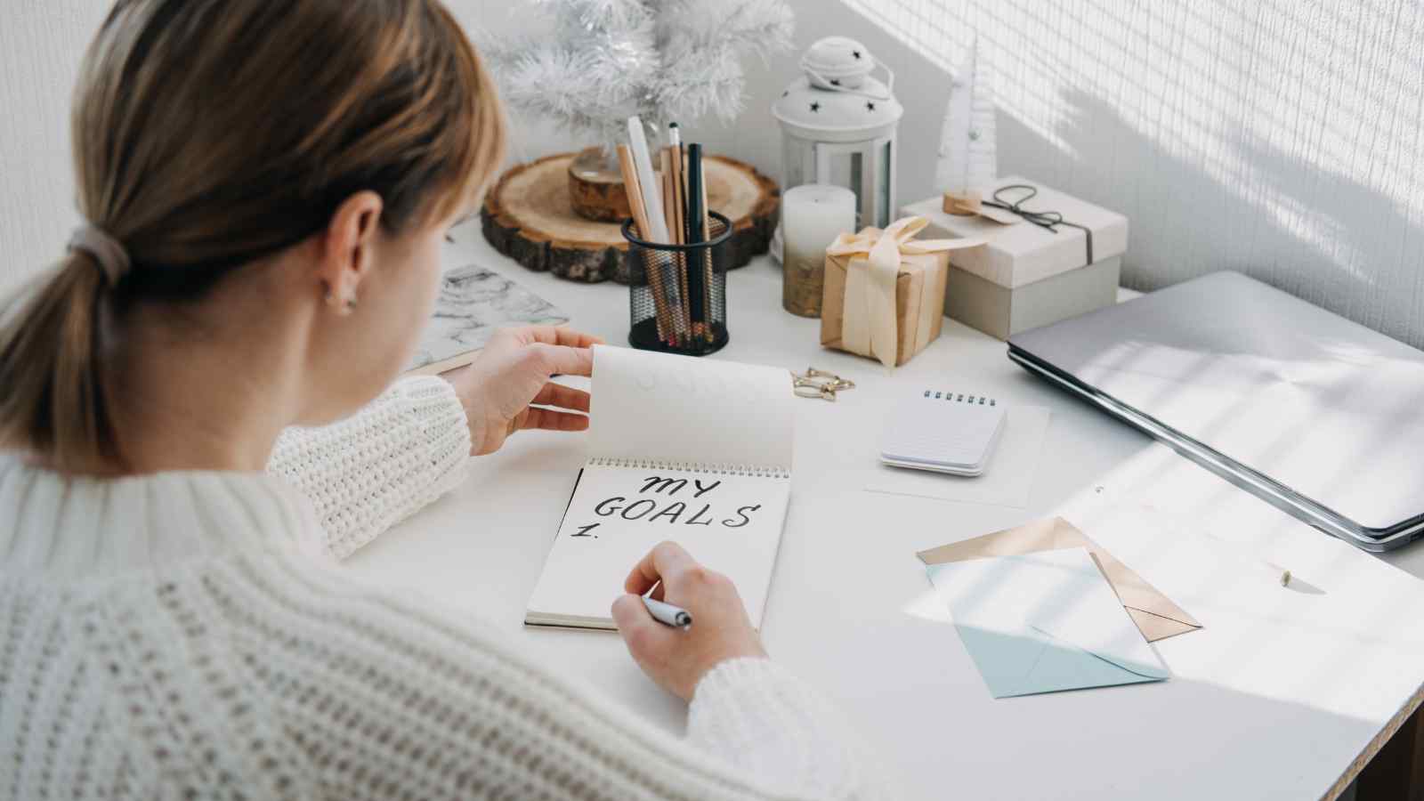 A person sitting at a desk writes "My Goals" in a notebook. The desk has stationery, a laptop, and decorative items.