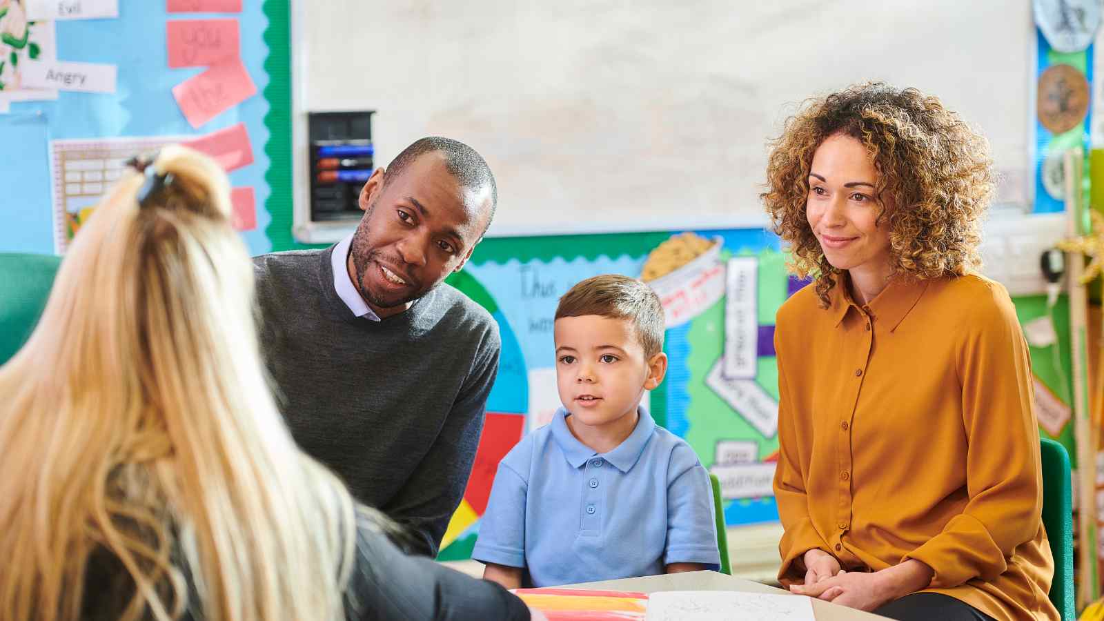Three adults and one child sit together in a classroom setting, with educational posters on the walls. The child is wearing a blue shirt, and they appear to be attending a meeting or discussion.
