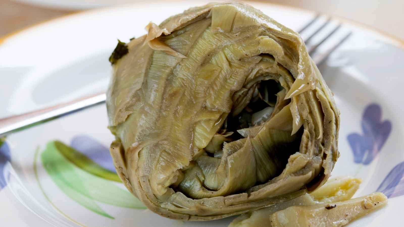 Close-up of a cooked artichoke on a decorative plate with a fork beside it, highlighting its layered structure.