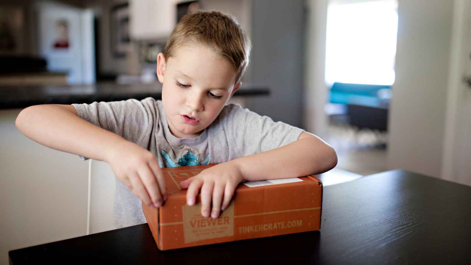 A young boy sits at a table, focused on opening a cardboard box with his hands in a home setting.