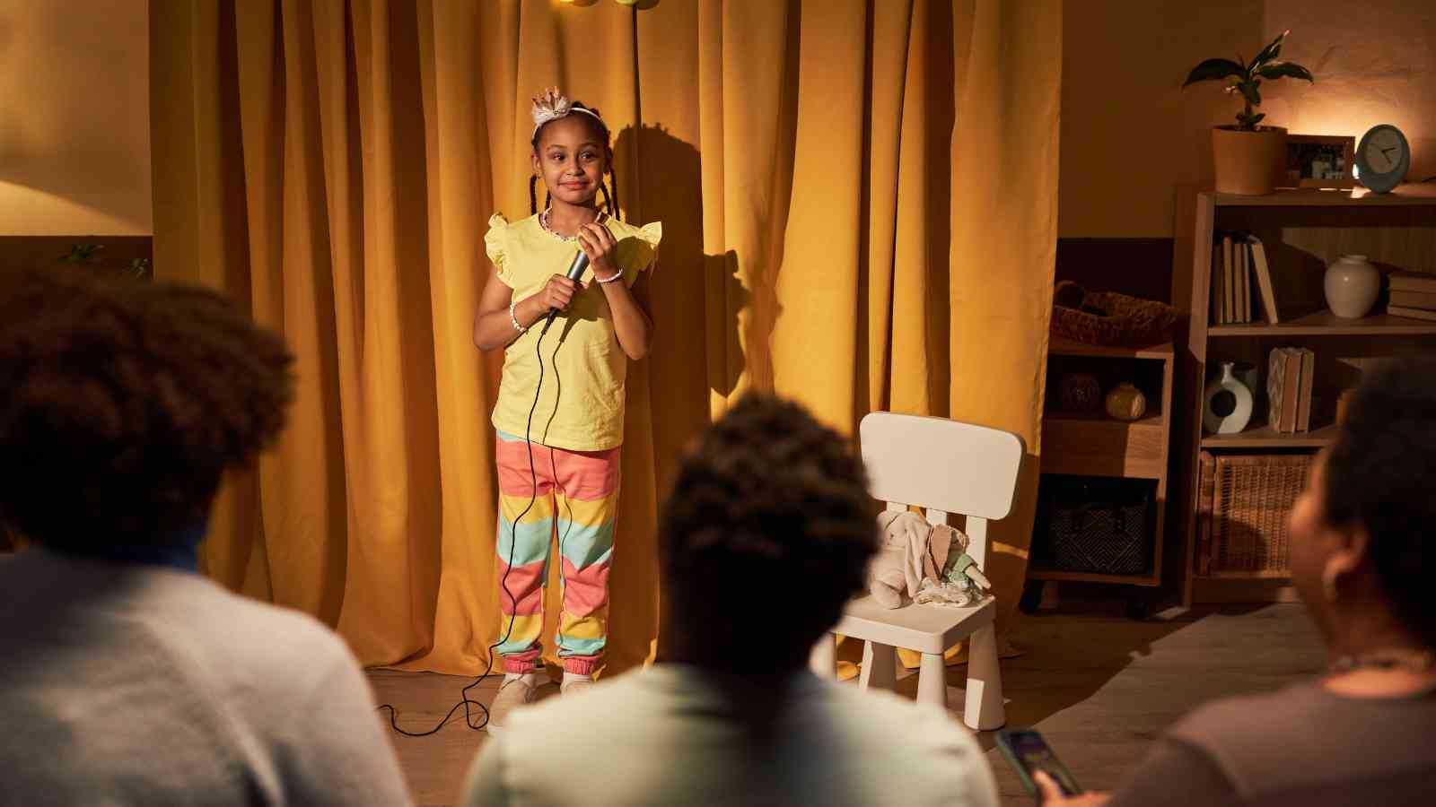 A child in colorful pants and a crown stands on a stage with a microphone, facing an audience seated in a room with yellow curtains and shelves.