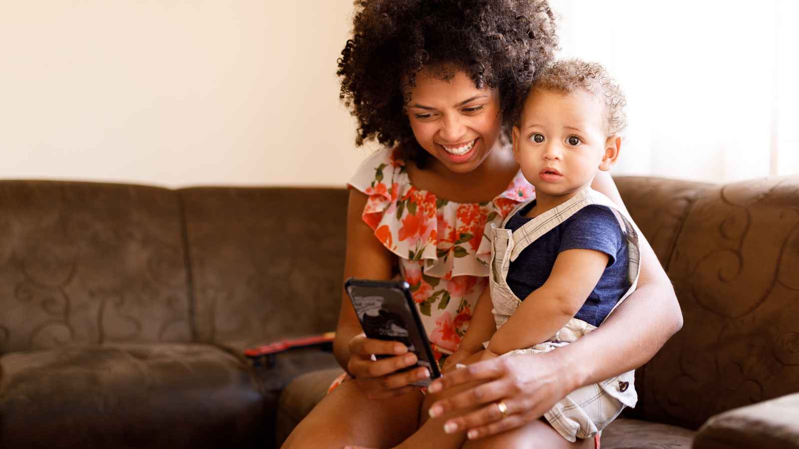 A woman smiles at her phone while holding a child on a sofa. The woman wears a floral top, and the child is dressed in a navy shirt and beige overalls.