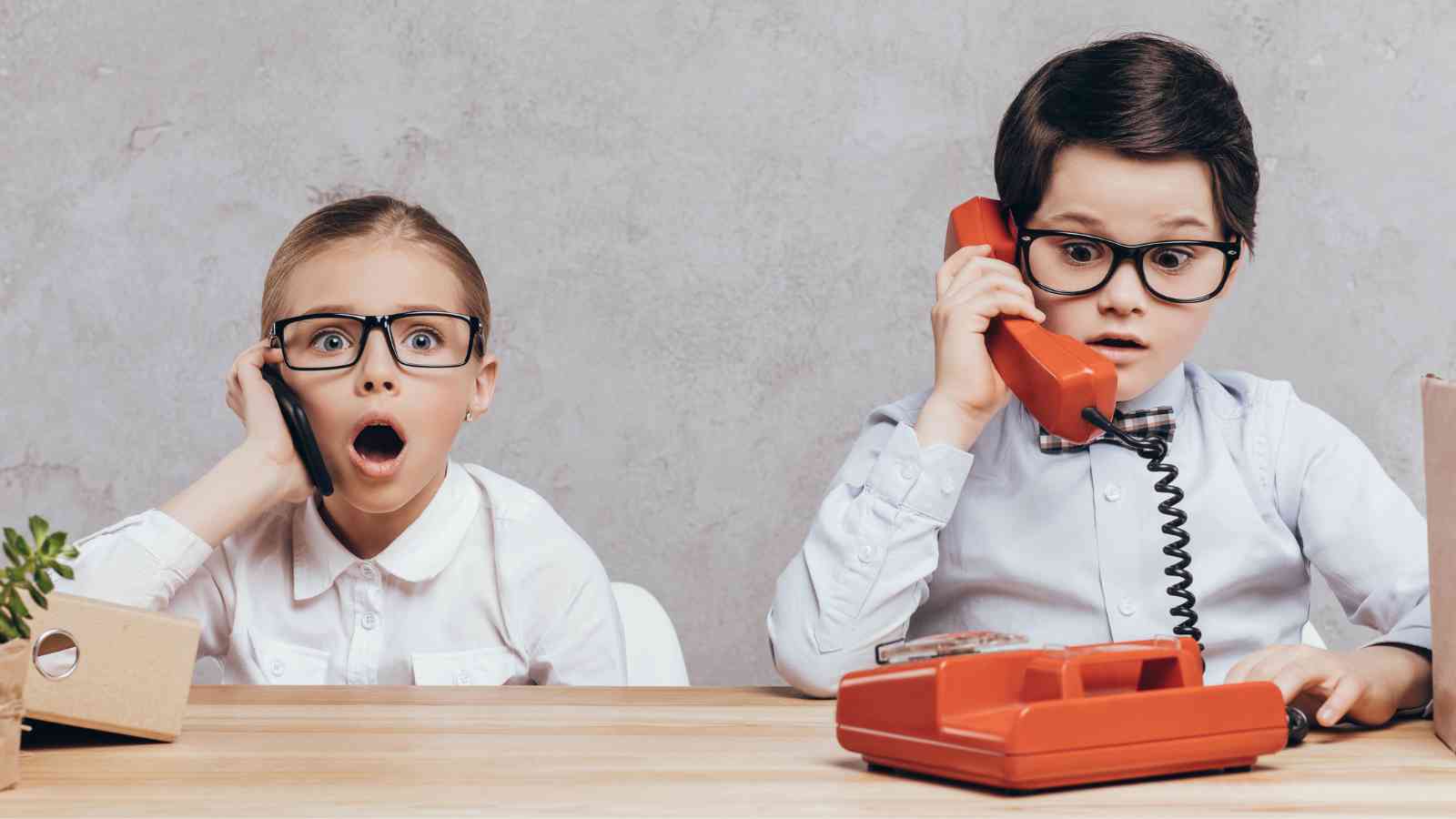 Two children in glasses at a desk using vintage phones. The girl appears shocked, while the boy looks surprised. A small plant and a folder are on the wooden table.