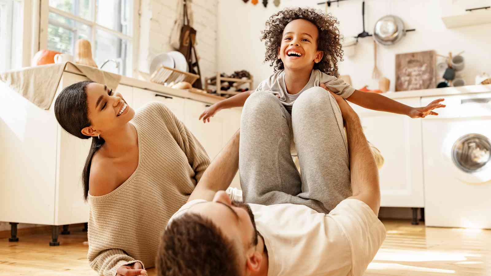 A child is playfully lifted by an adult lying on the floor, while another adult smiles nearby in a bright kitchen.