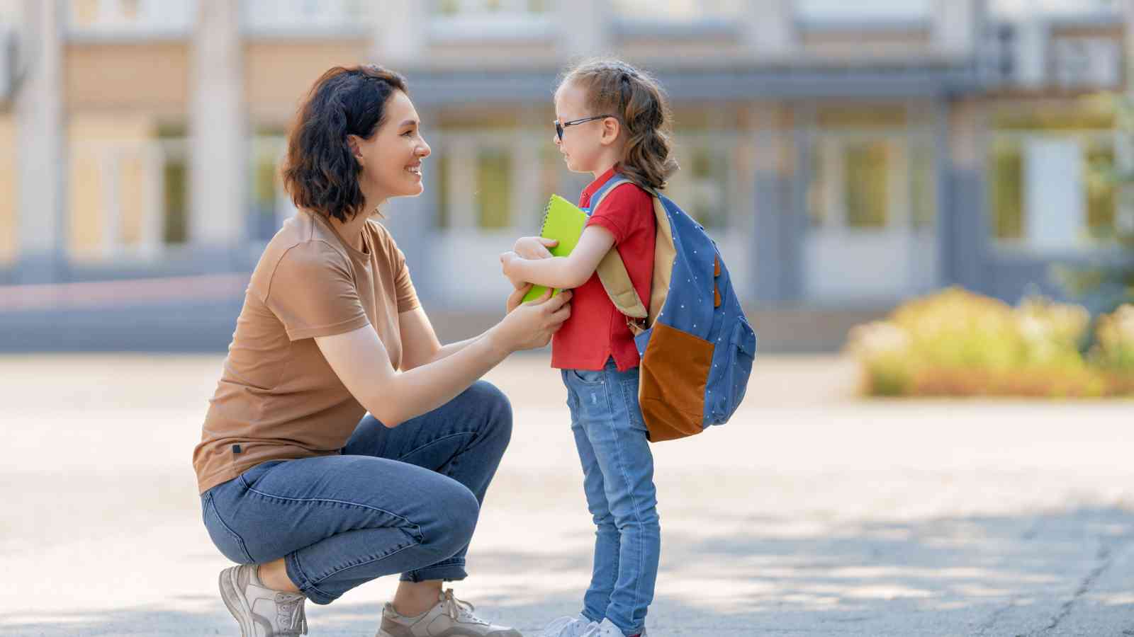An adult woman crouches and smiles at a young girl with a backpack and notebook, standing outdoors in front of a building.