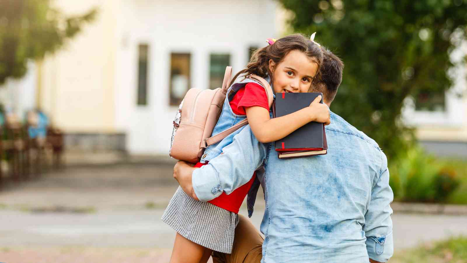 A man carries a young girl wearing a backpack and holding books outdoors. The girl looks over his shoulder, facing the camera.