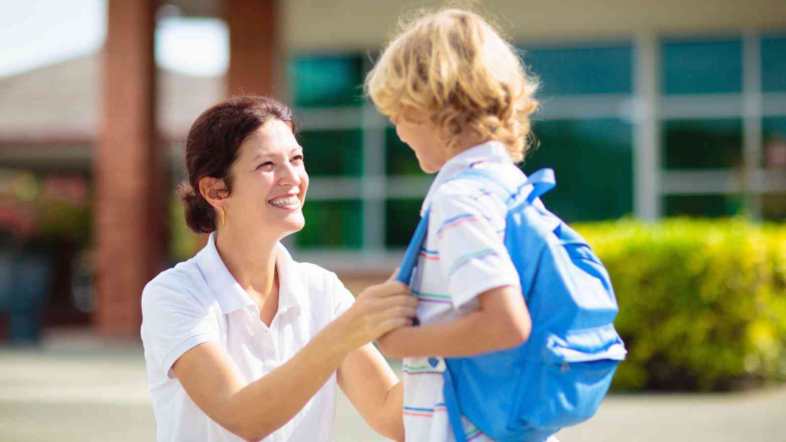 An adult woman kneels and smiles at a young boy wearing a blue backpack outside, possibly at a school or daycare.