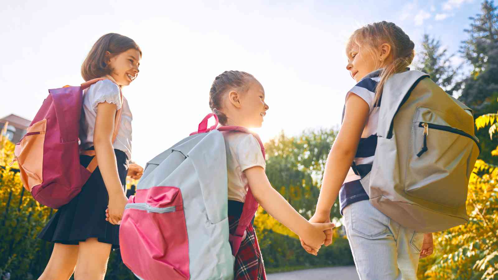 Three young girls with backpacks walk outdoors, holding hands and smiling at each other on a sunny day.