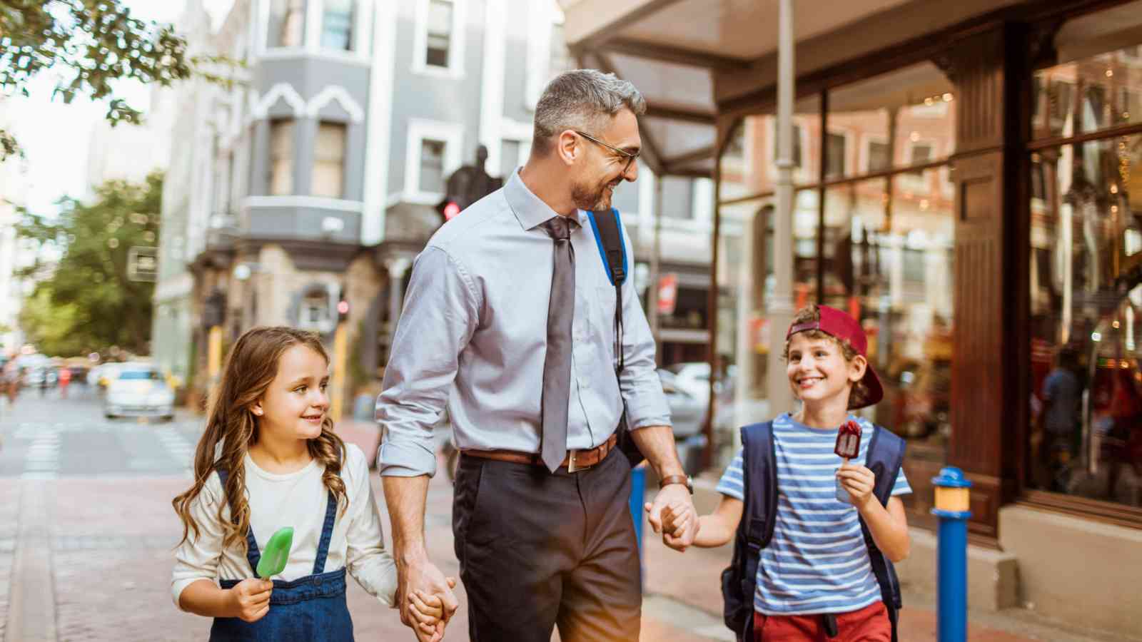 A man in business attire walks down a city street holding hands with a young girl and boy, who are eating ice cream and smiling.