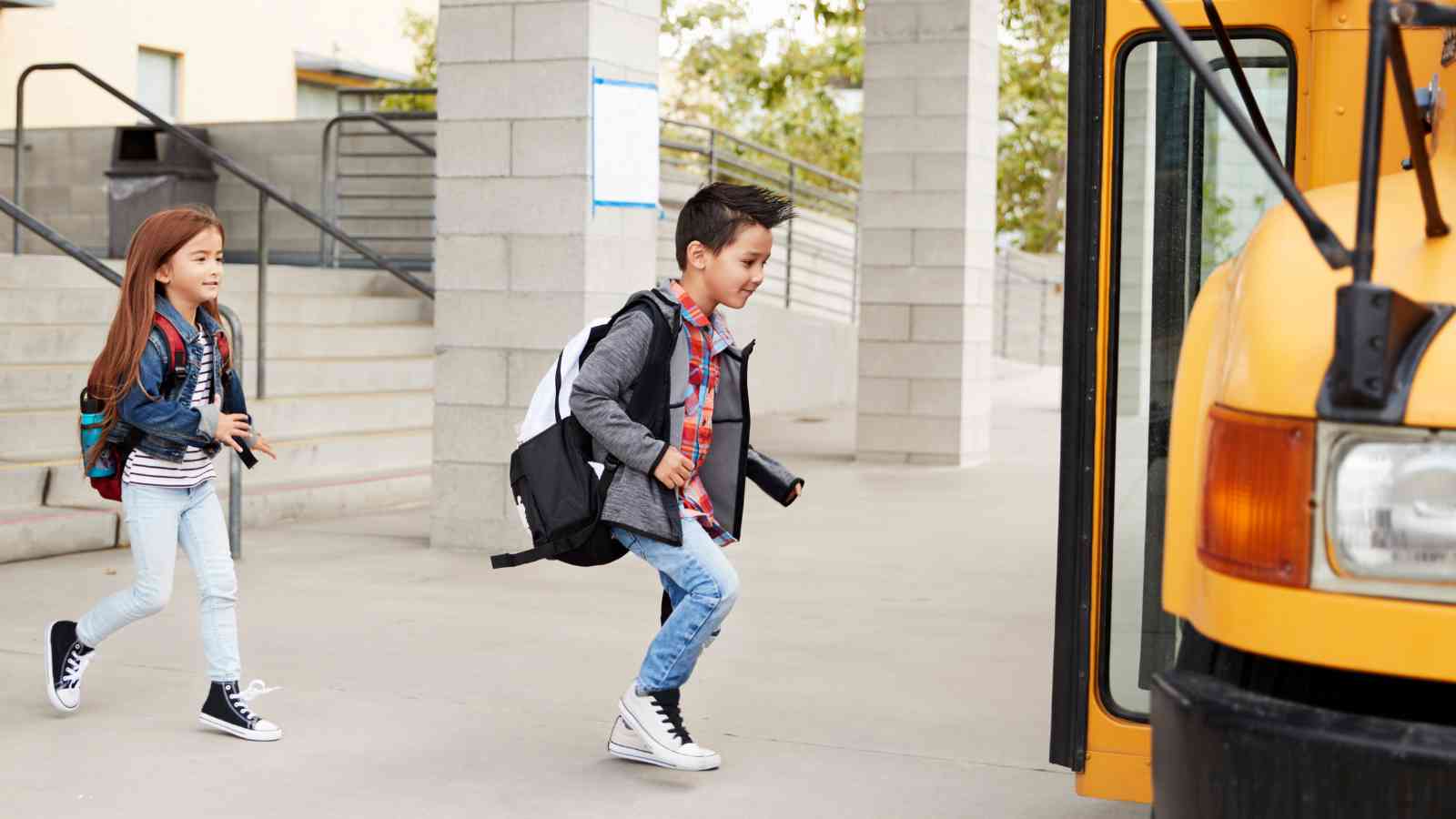 Two children with backpacks walk quickly toward the open door of a yellow school bus parked at a school.