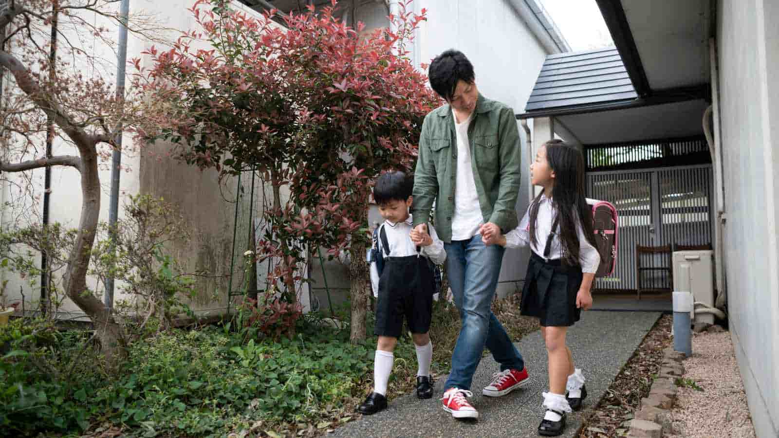 An adult walks outside with a young boy and girl in school uniforms, holding their hands.