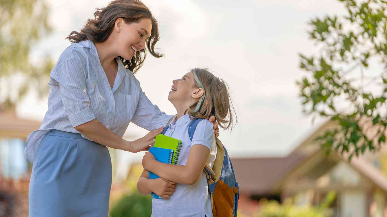 An adult woman smiles and talks to a young girl holding notebooks and wearing a backpack outdoors, suggesting a school setting.