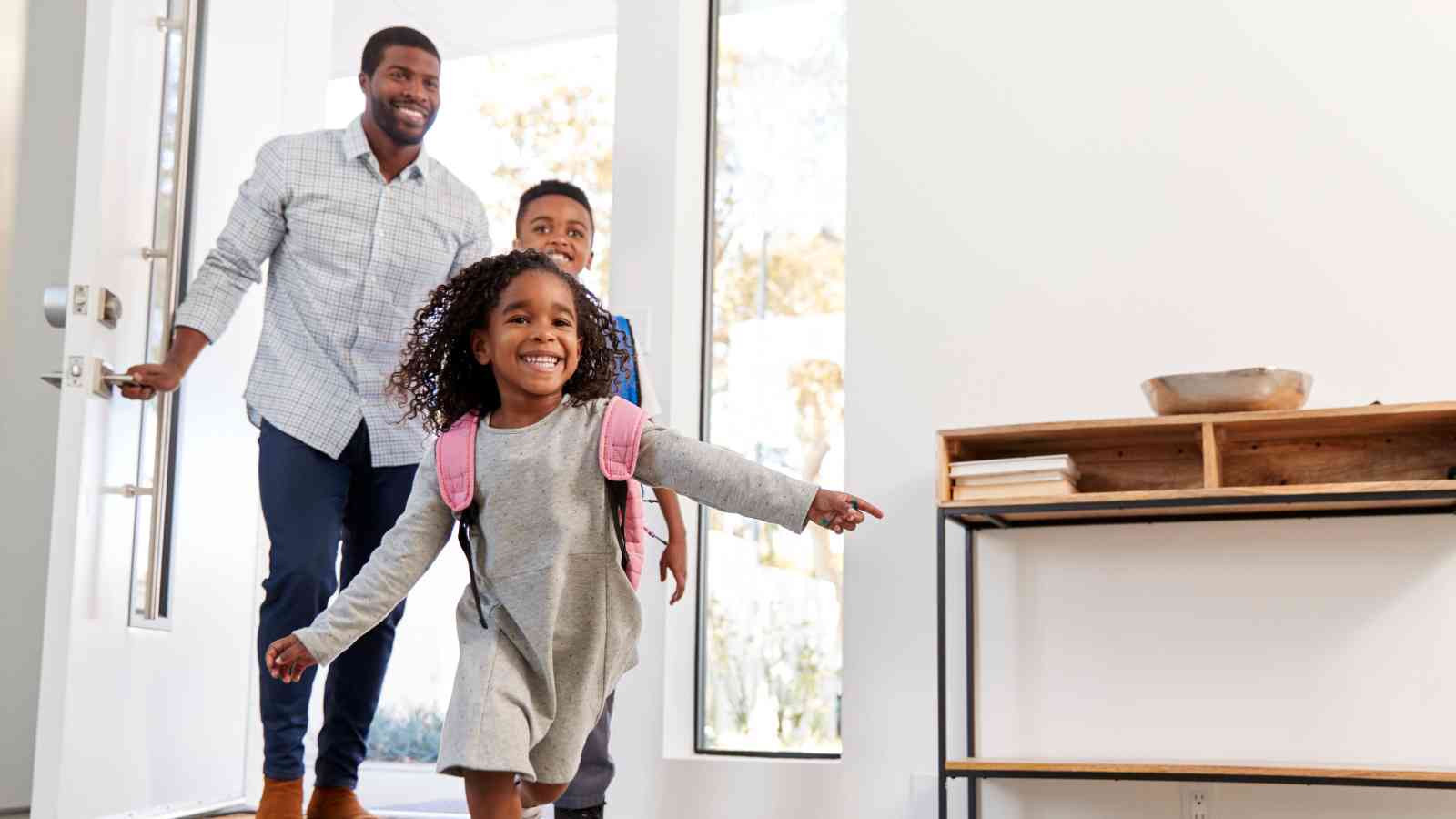 A man and two children enter a bright home, with the girl in front wearing a backpack and smiling while the boy and man follow behind.