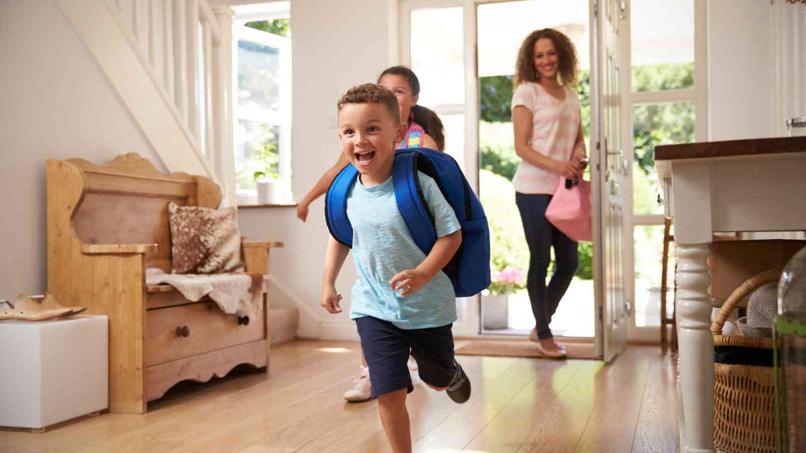 A young boy with a backpack runs excitedly into a house, followed by another child and a woman entering through the door.