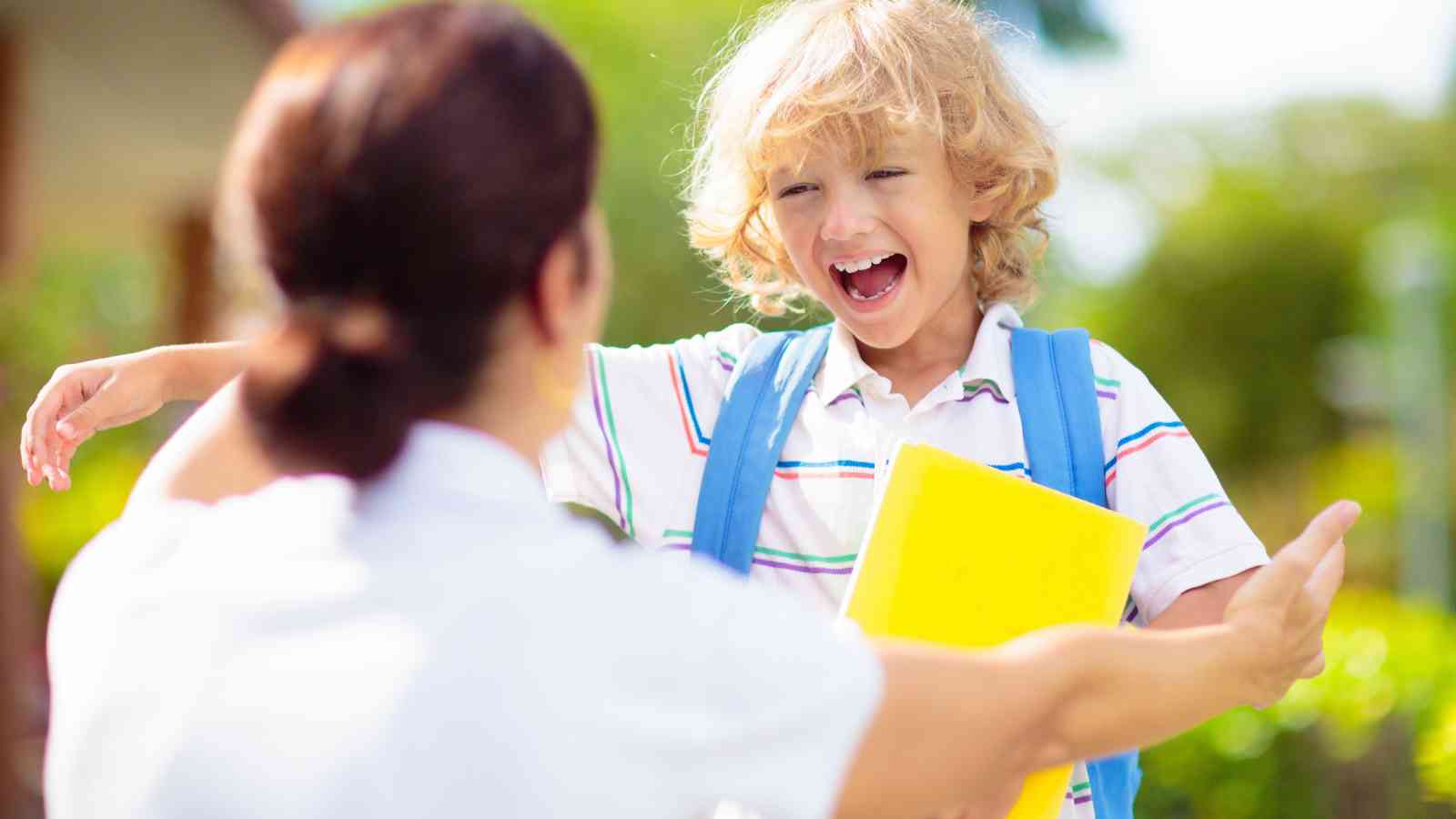 A young child with a backpack smiles and holds a yellow folder while reaching out to an adult with open arms outdoors.