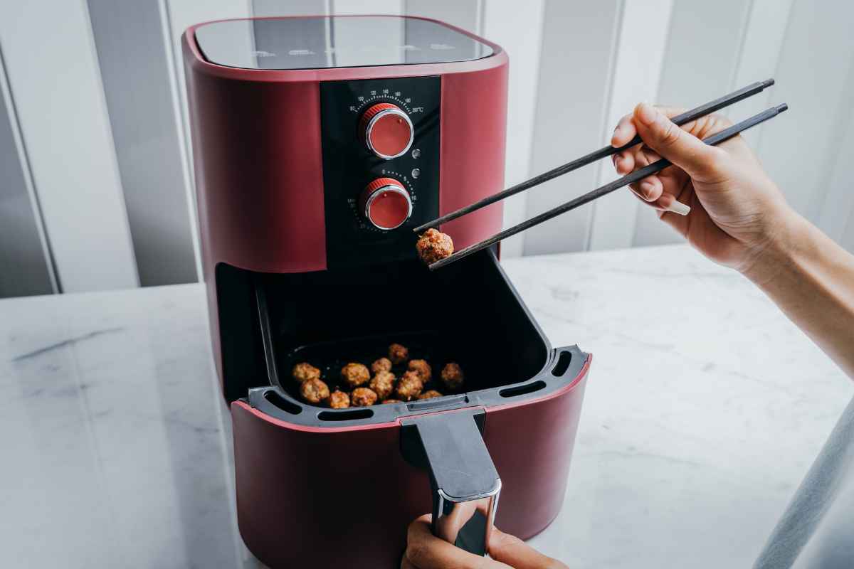 A person uses chopsticks to pick up a cooked meatball from the basket of a red air fryer on a white countertop.