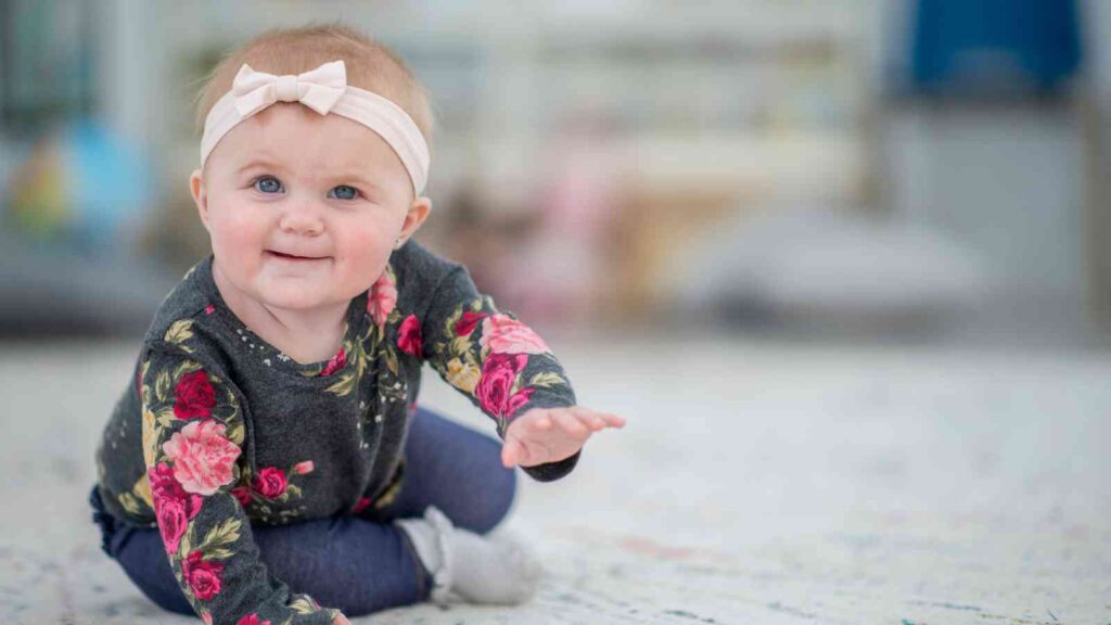 A baby wearing a floral shirt, pink headband, and jeans is sitting on a patterned floor, reaching forward with one hand.