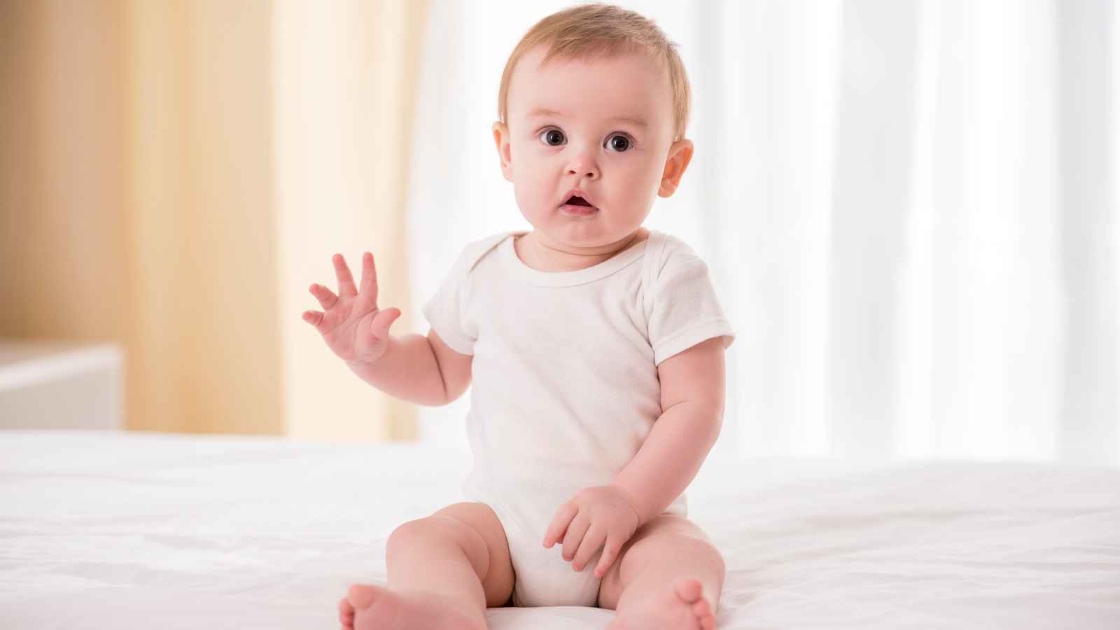 Baby boy wearing a white onesie, sitting in a bed behind a white curtain as background.