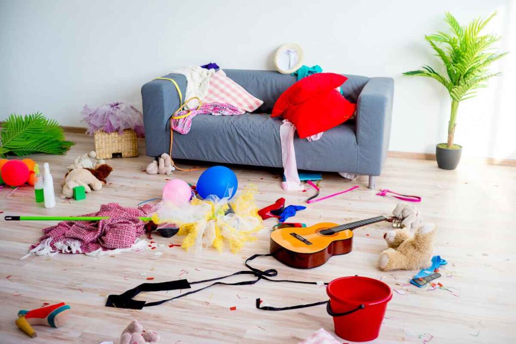 A messy living room with toys, clothes, a guitar, pillows, and various items scattered on the floor in front of a gray sofa and a potted plant.