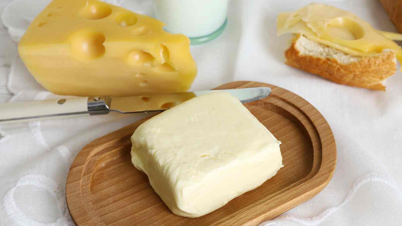 Butter on a wooden tray, a knife, a wedge of cheese, a glass of milk, and a slice of bread with cheese on a cloth.