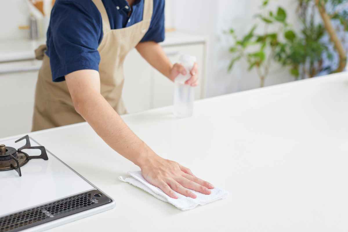A person wearing an apron wipes a kitchen counter with a cloth while holding a spray bottle.