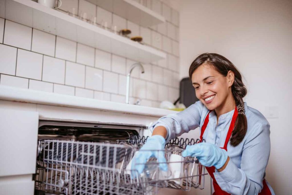 A woman wearing blue gloves and a red apron loads dishes into a dishwasher in a kitchen with white tile backsplash.