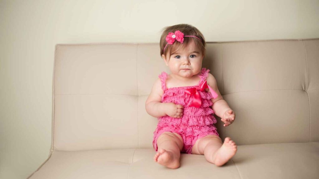 Baby wearing a pink ruffled outfit and headband sits on a beige couch against a plain wall.
