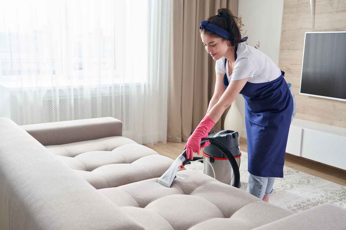 A woman in an apron and gloves is cleaning a beige sofa with a vacuum cleaner in a living room.