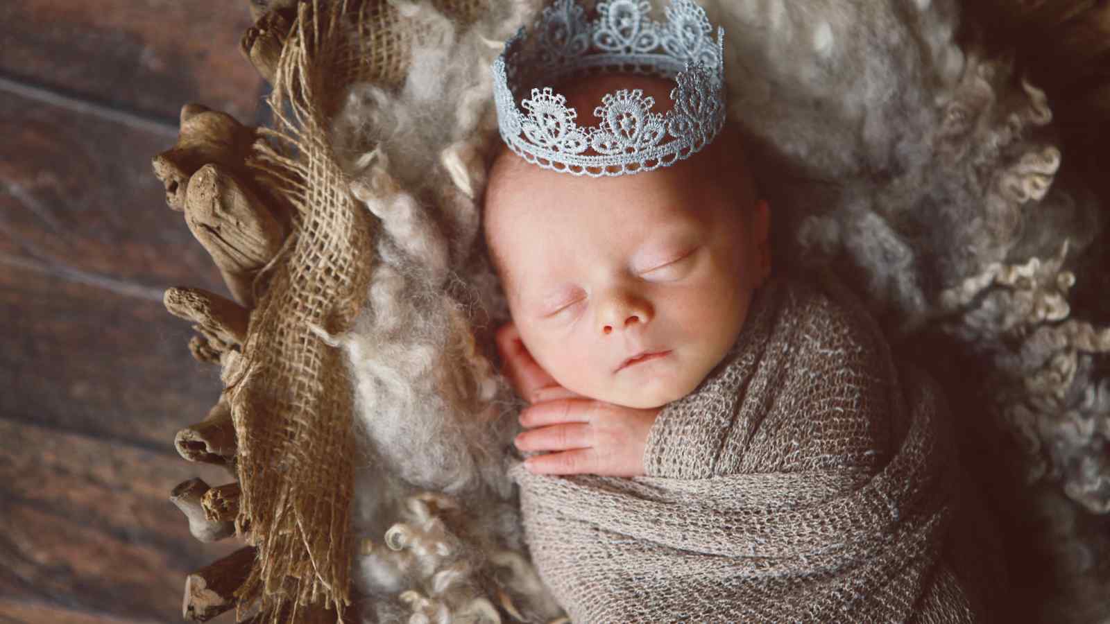 A newborn baby wrapped in a brown blanket sleeps on a soft, textured surface while wearing a small silver crown.