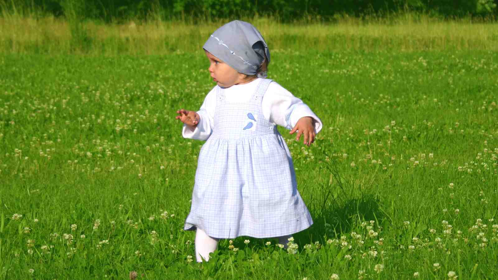 A young child in a white dress and gray headscarf, stands in a grassy field dotted with small white flowers.