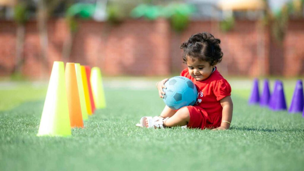 A young child in a red outfit sits on grass holding a blue soccer ball, with colorful cones lined up nearby.