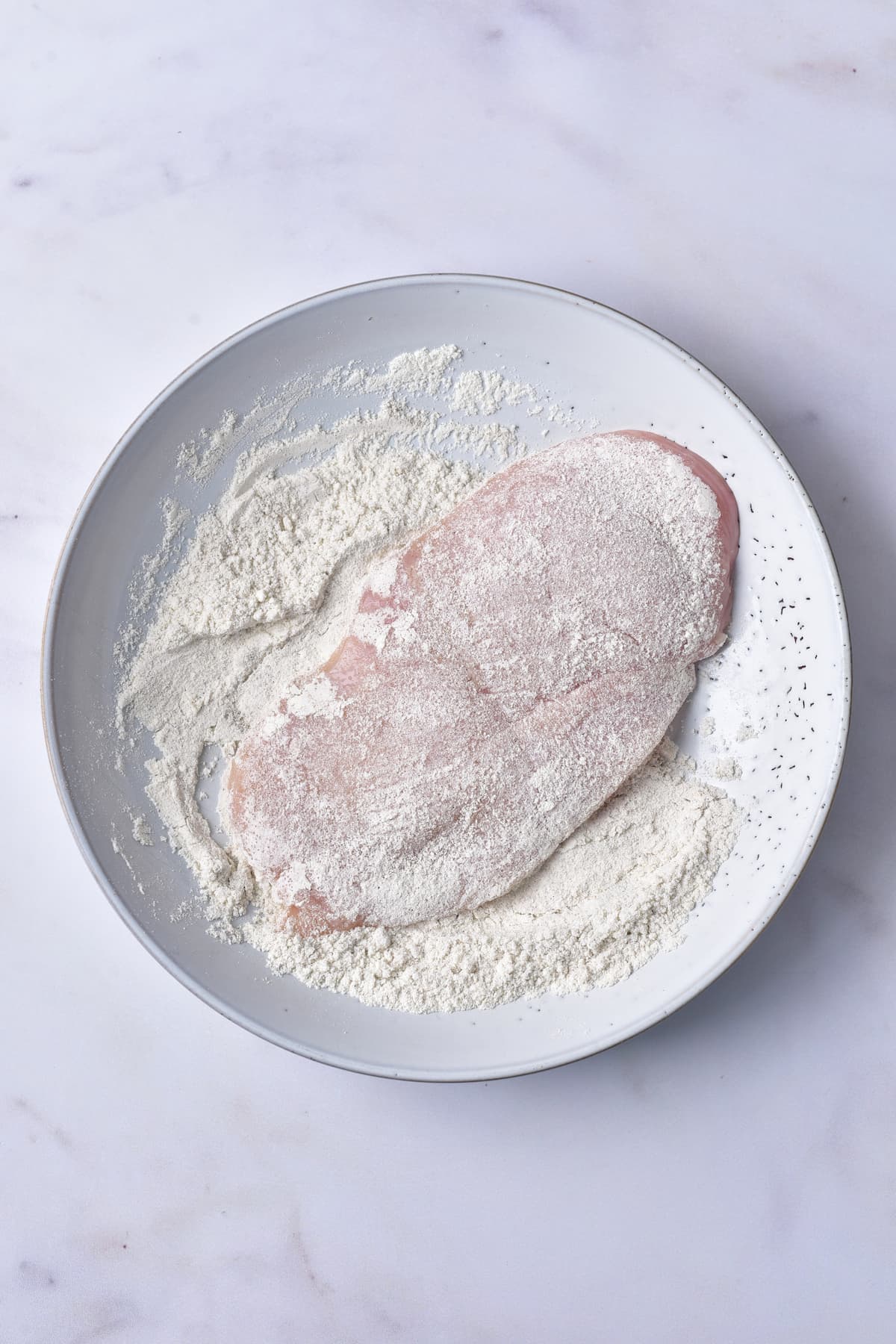 A raw chicken breast coated in flour on a white plate, placed on a light-colored marble surface.