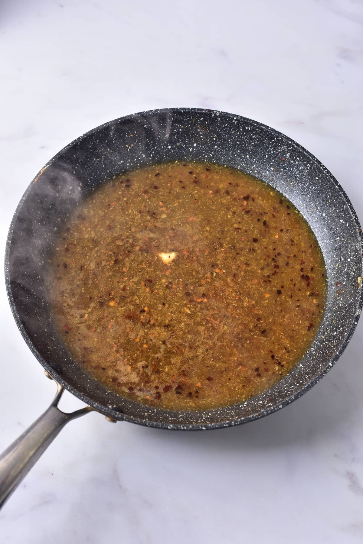 A black frying pan on a white surface contains a simmering brown sauce with visible herbs and spices. Steam rises gently from the flavorful mixture.