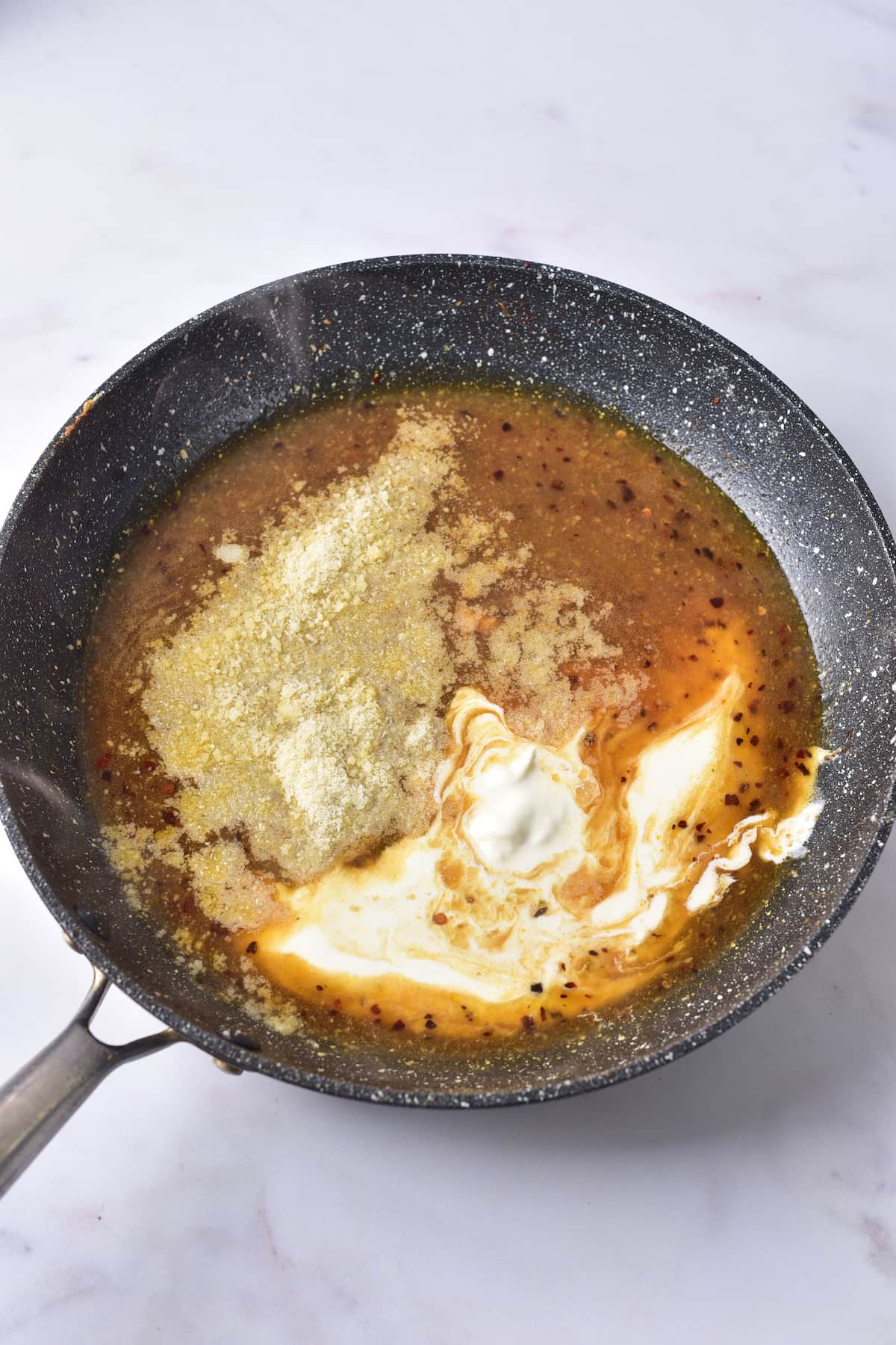 A nonstick frying pan with savory marry me chicken in brown sauce, topped with grated cheese and a dollop of cream, on a white surface.