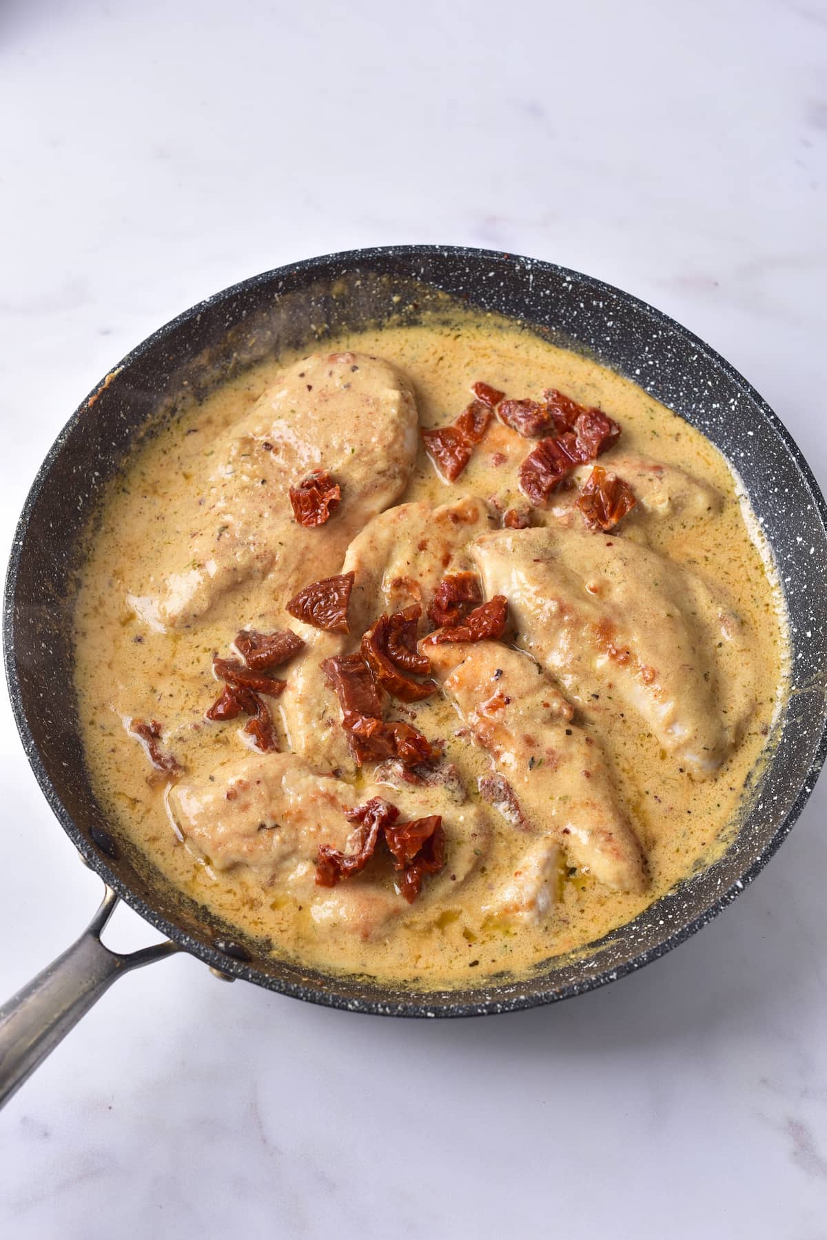 A black pan containing marry me chicken—tender chicken breasts in a creamy sauce, topped with pieces of sun-dried tomatoes—on a white surface.