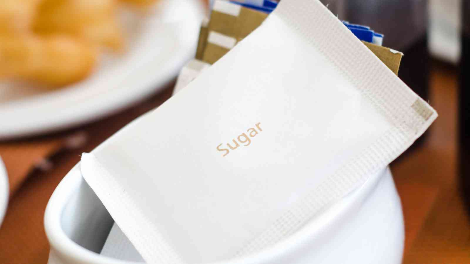 A close-up of a white packet labeled "Sugar" placed inside a white container with other sweetener packets in the background.