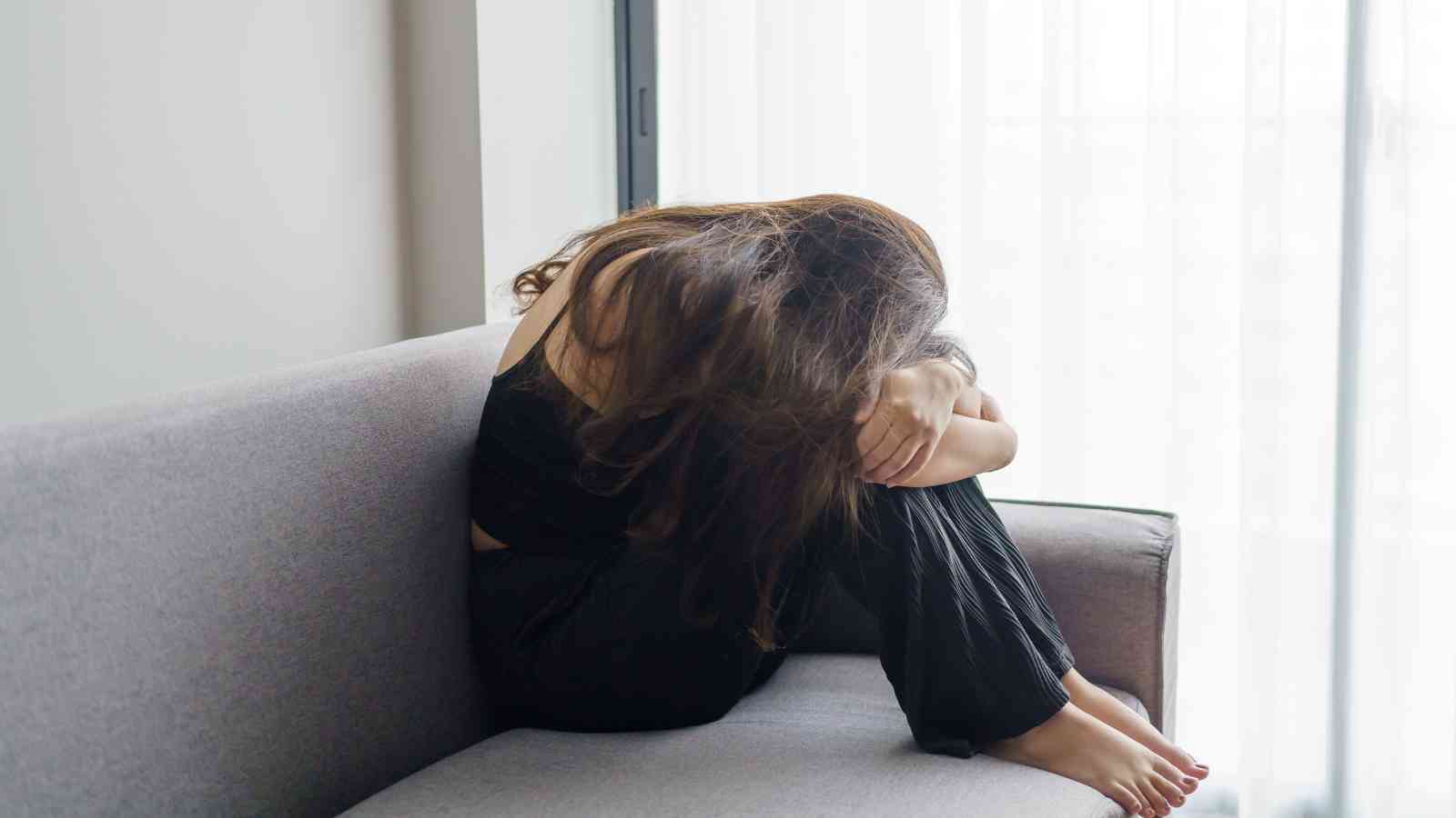 A person sits on a gray couch with knees drawn to chest, head down, and arms wrapped around legs, appearing distressed.