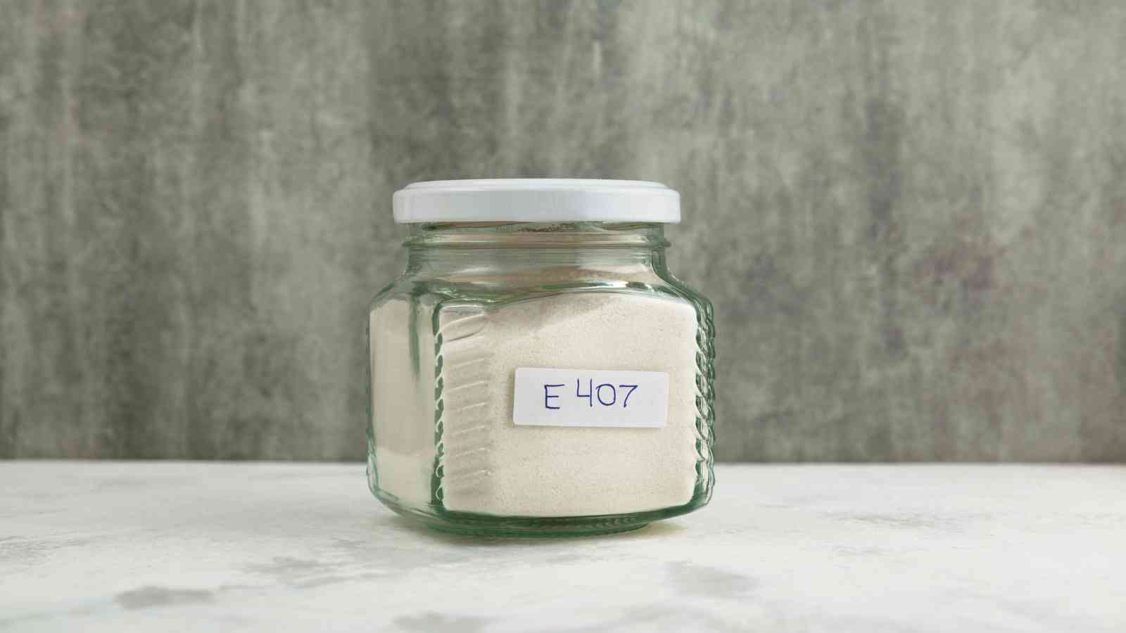 A glass jar with a white lid containing a white powder labeled "E 407" sits on a light surface against a gray background.