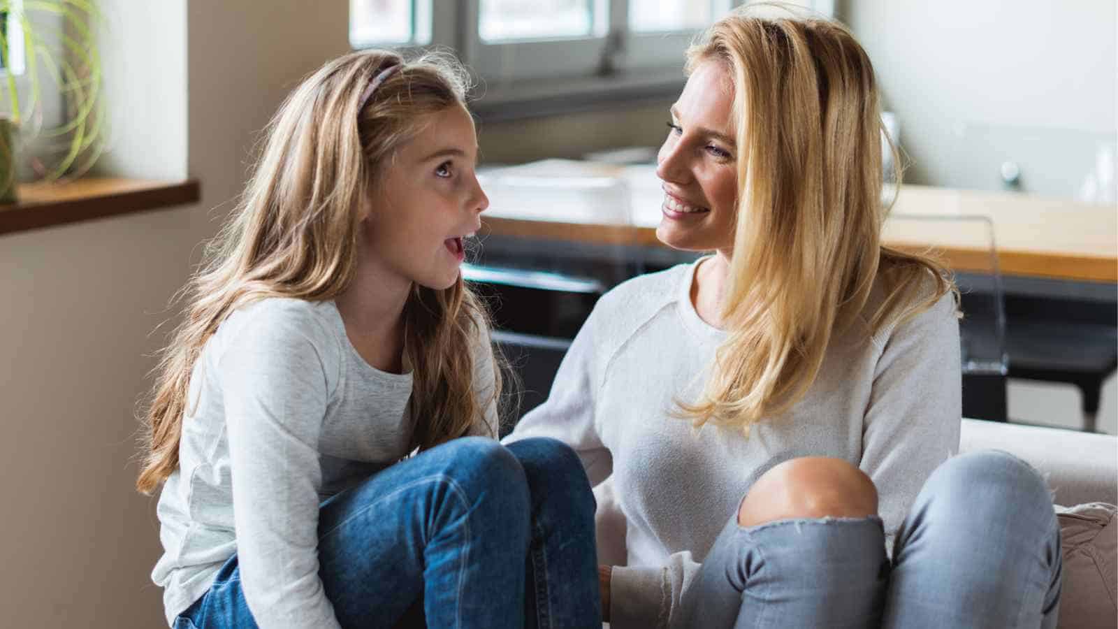 A woman and a young girl sit on a couch indoors, facing each other and talking, both wearing casual clothes.