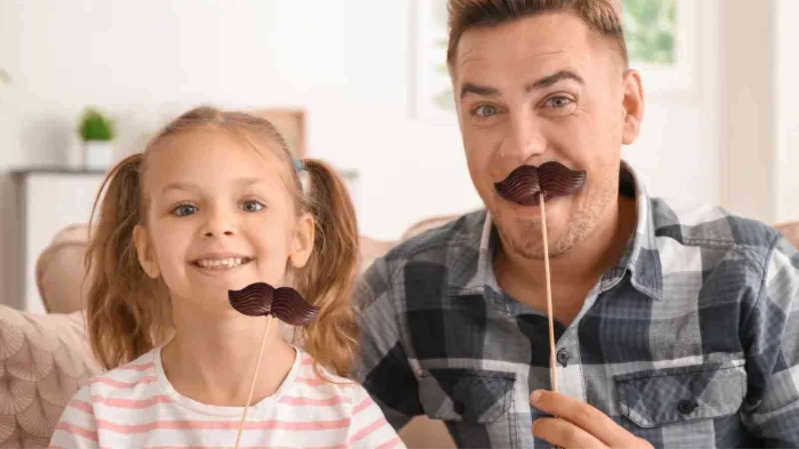 A man and a young girl sit together holding fake mustaches on sticks in front of their faces, smiling at the camera.