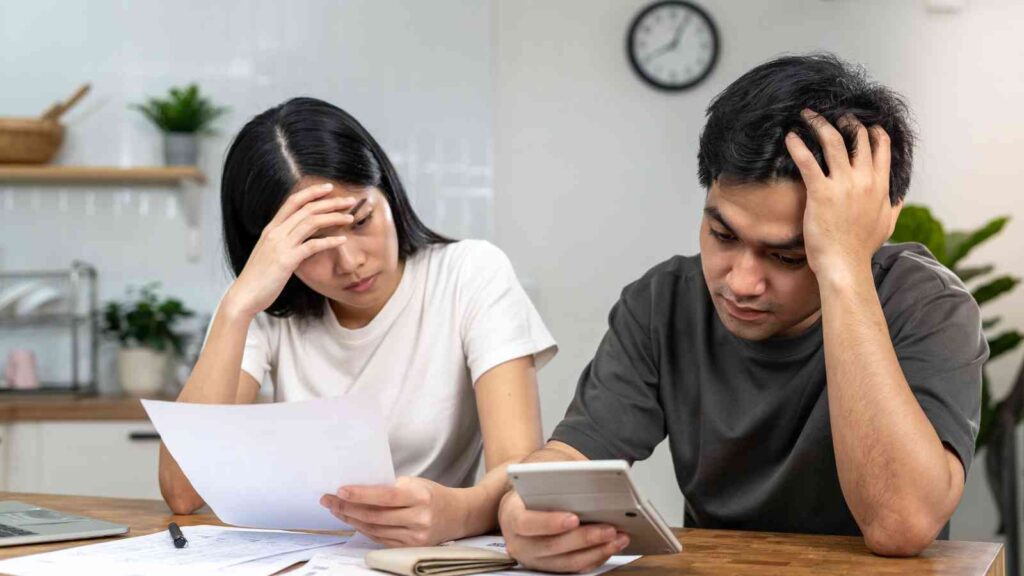A man and woman sit at a table looking stressed while reviewing papers and using a calculator, with a clock on the wall behind them.