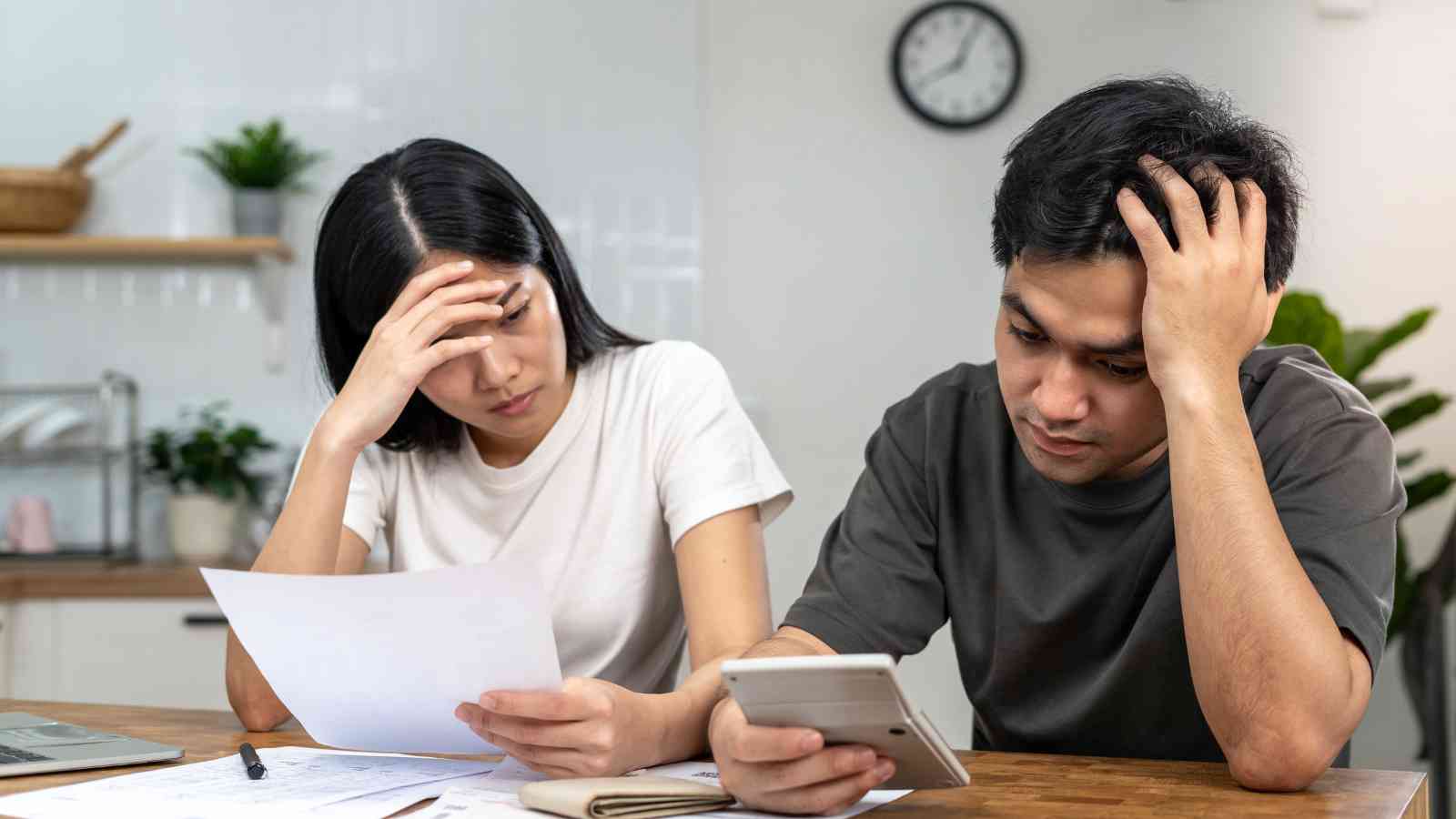 A man and woman sit at a table looking stressed while reviewing papers and using a calculator, with a clock on the wall behind them.