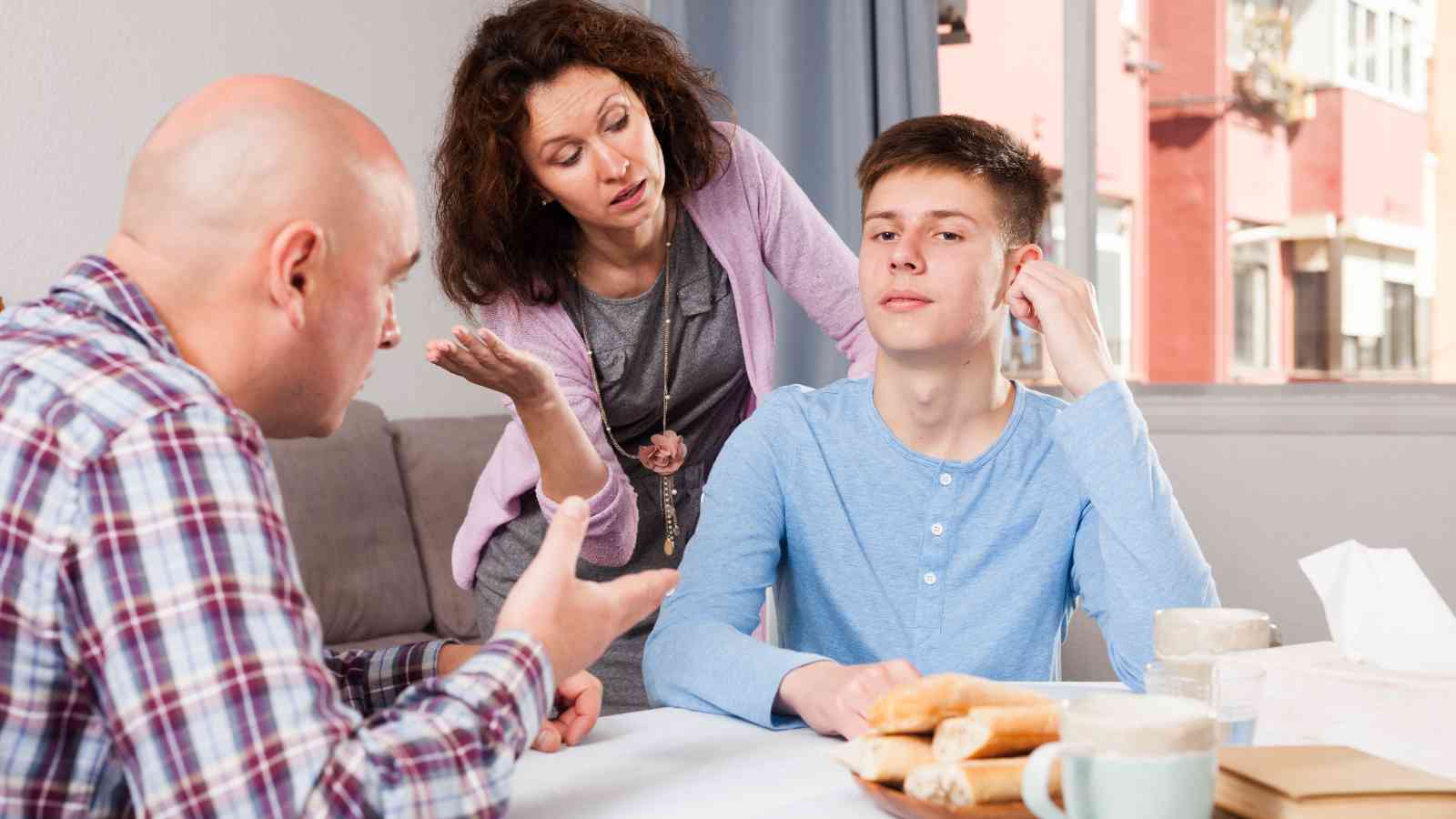 A teenage boy sits at a table with food, looking away as a man and woman, possibly his parents, talk to him with concerned and frustrated expressions.