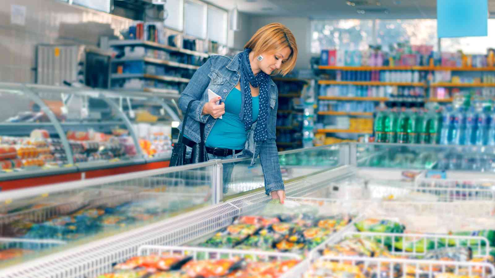 A woman in a blue outfit shops for frozen food in a supermarket, reaching into a freezer aisle while holding a shopping list.