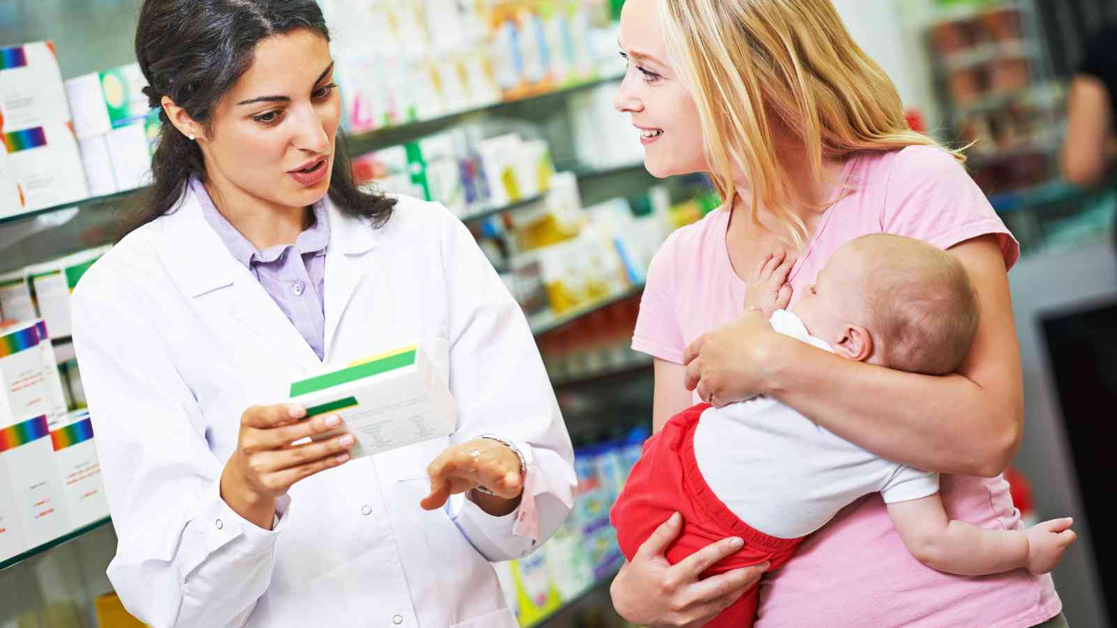 A pharmacist explains medication details to a woman holding a baby inside a pharmacy. Shelves with various products are visible in the background.