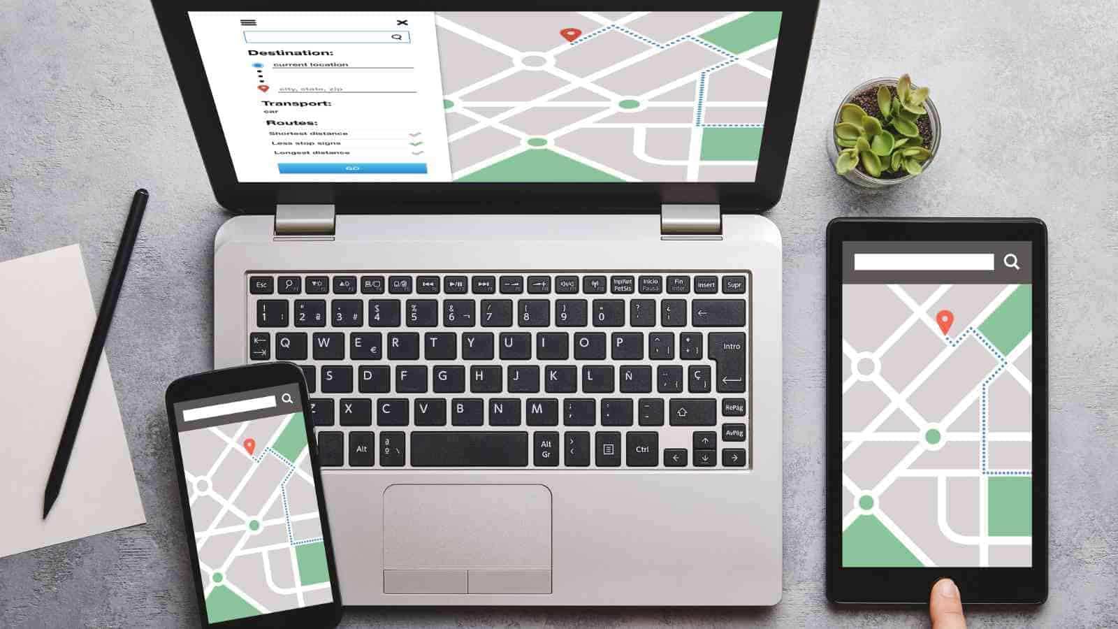 A laptop, smartphone, and tablet display digital maps with location pins on a gray surface next to a small plant and notepad.